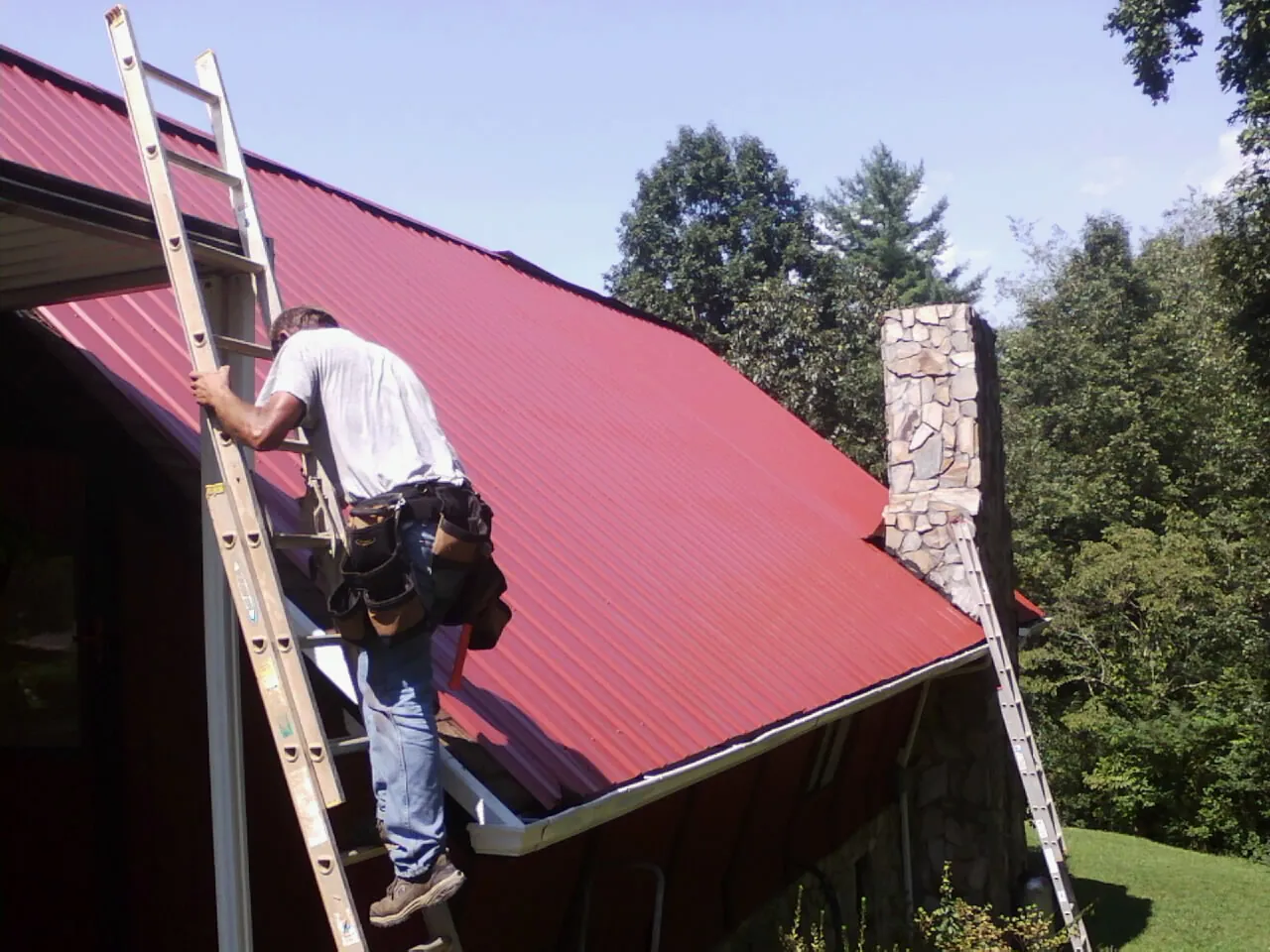 Roofer on a ladder beside a red metal roof and stone chimney — Soffit & Fascia Repair by Roofing Brothers in Kaleva